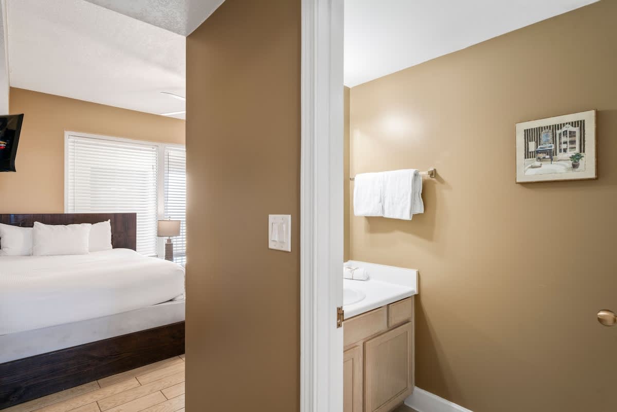 Partial view of a bathroom with a single vanity featuring a white countertop and light wood cabinets, a mirror, and a towel rack, with a bedroom visible through the doorway.