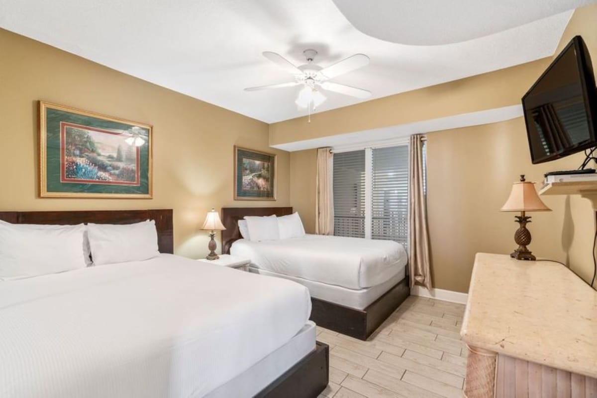 Bright bedroom featuring a queen bed and a twin bed with white linens and dark wood frames, light wood-look tile flooring, a ceiling fan, and a sliding glass door leading to an outdoor space.