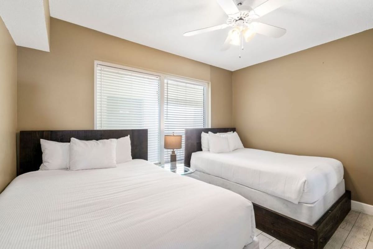 Bedroom with a queen bed and a twin bed, both with dark wood headboards and white bedding, a ceiling fan, and a large window with white blinds.