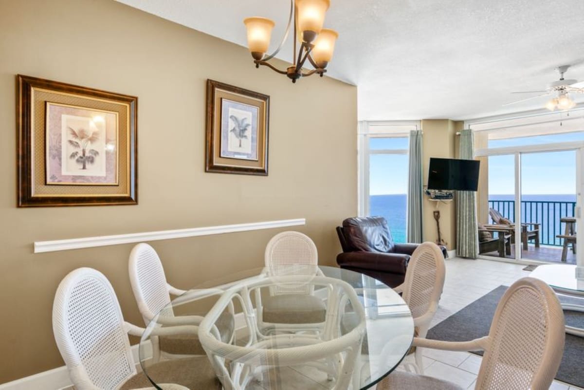 Bright dining space with a round glass table and white wicker chairs, featuring warm beige walls adorned with framed botanical prints, and offering a glimpse of the ocean through large windows.