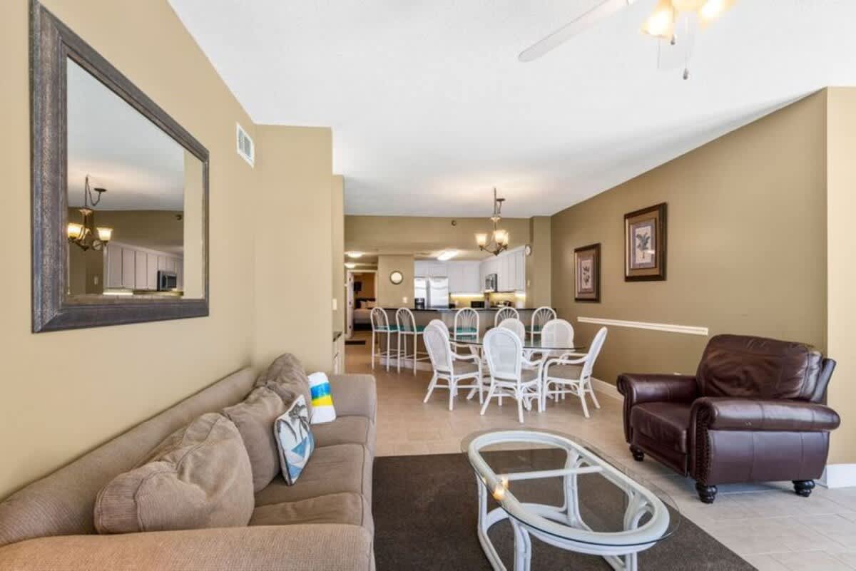 Expansive living room with a comfortable sofa and leather armchair, a glass coffee table, and a large wall mirror, seamlessly connecting to the dining area and kitchen, all set on light tile flooring.