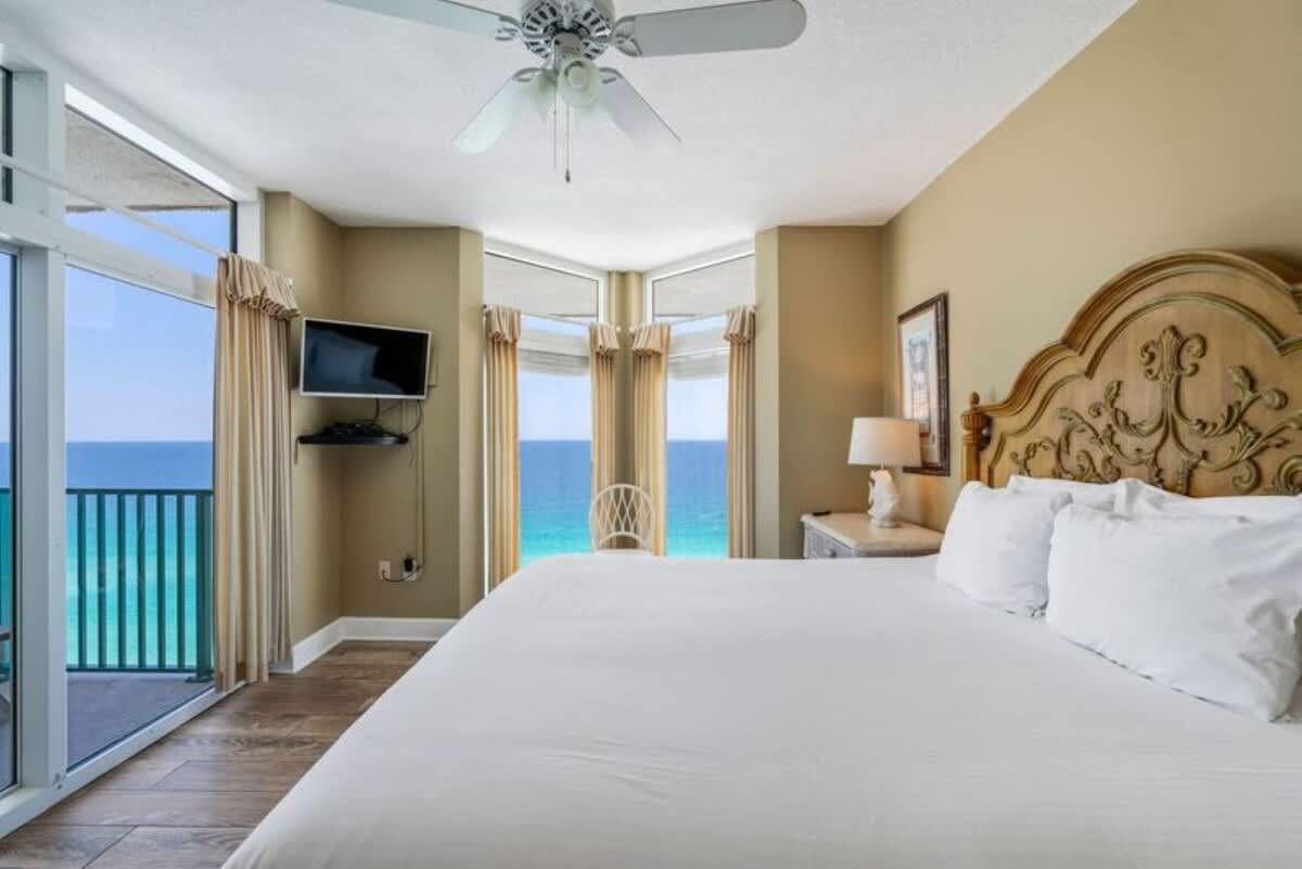Master bedroom featuring a king-sized bed with a decorative wooden headboard, wood-look flooring, and expansive windows with ocean views. A wall-mounted TV and a small white chair are visible near the windows.