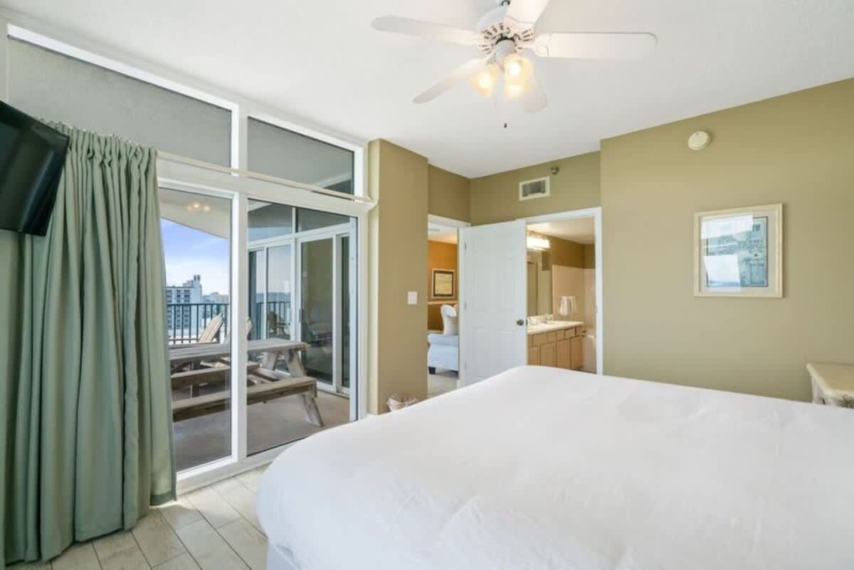 The master bedroom features a white king bed and access to a private balcony with a picnic table, offering city and partial ocean views. An open doorway leads to an en-suite bathroom.