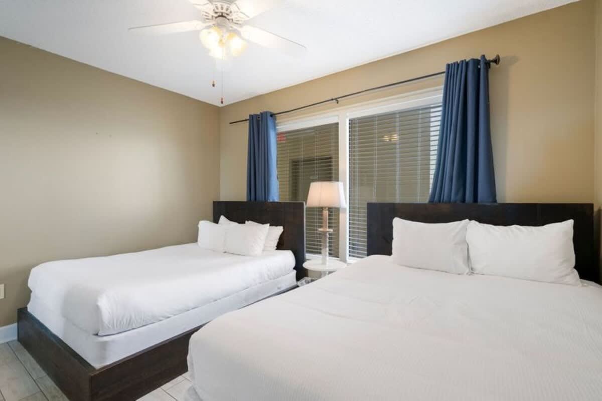 This bedroom contains two beds, one queen and one full, with dark wood frames and white bedding, separated by a small nightstand with a lamp. Blue curtains frame the window.