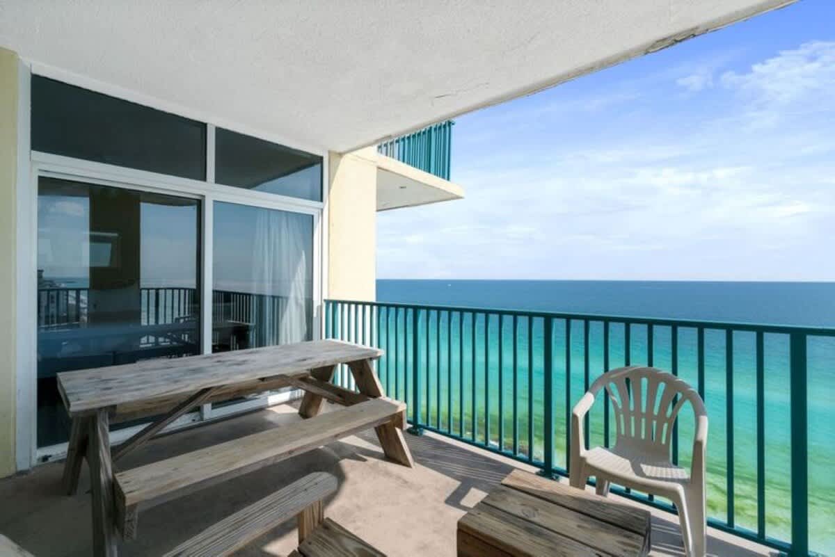 A spacious outdoor balcony overlooking the ocean, furnished with a rustic wooden picnic table and a white plastic chair. The turquoise railing provides a vibrant contrast against the deep blue sea and sky.