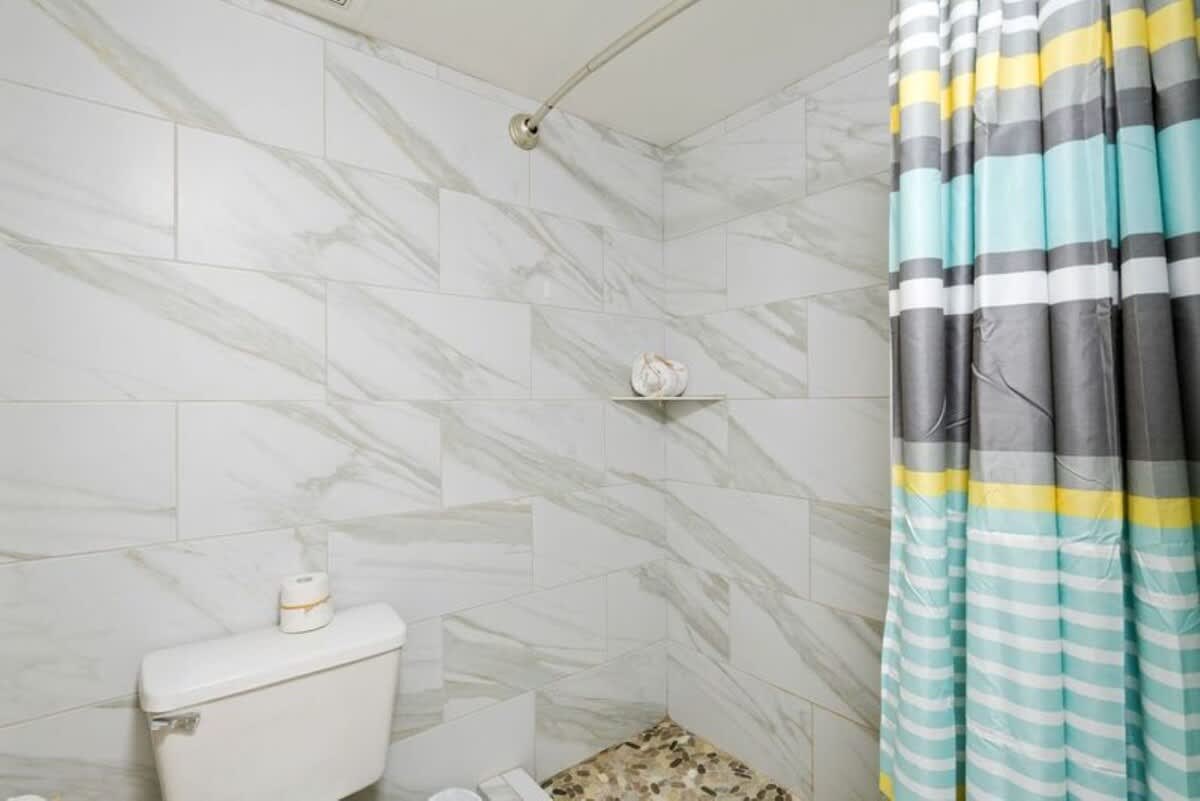 A contemporary shower stall with white marble-patterned rectangular tiles and a colorful striped shower curtain in teal, yellow, and gray. A white toilet is visible next to the shower, and the floor has small pebble-like tiles.