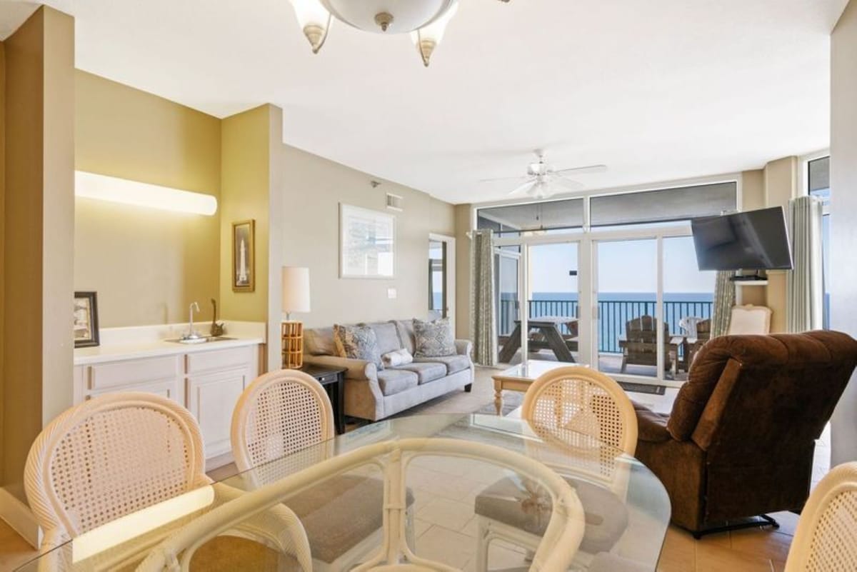 A view from the dining area shows the glass-top table and wicker chairs, with a wet bar to the left and the living room with ocean views visible through large windows in the background.