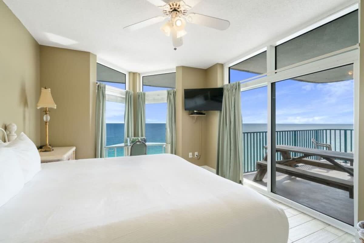 This master bedroom offers panoramic ocean views through floor-to-ceiling windows, with a white king bed and a small desk area. A wall-mounted TV and a ceiling fan are also visible.