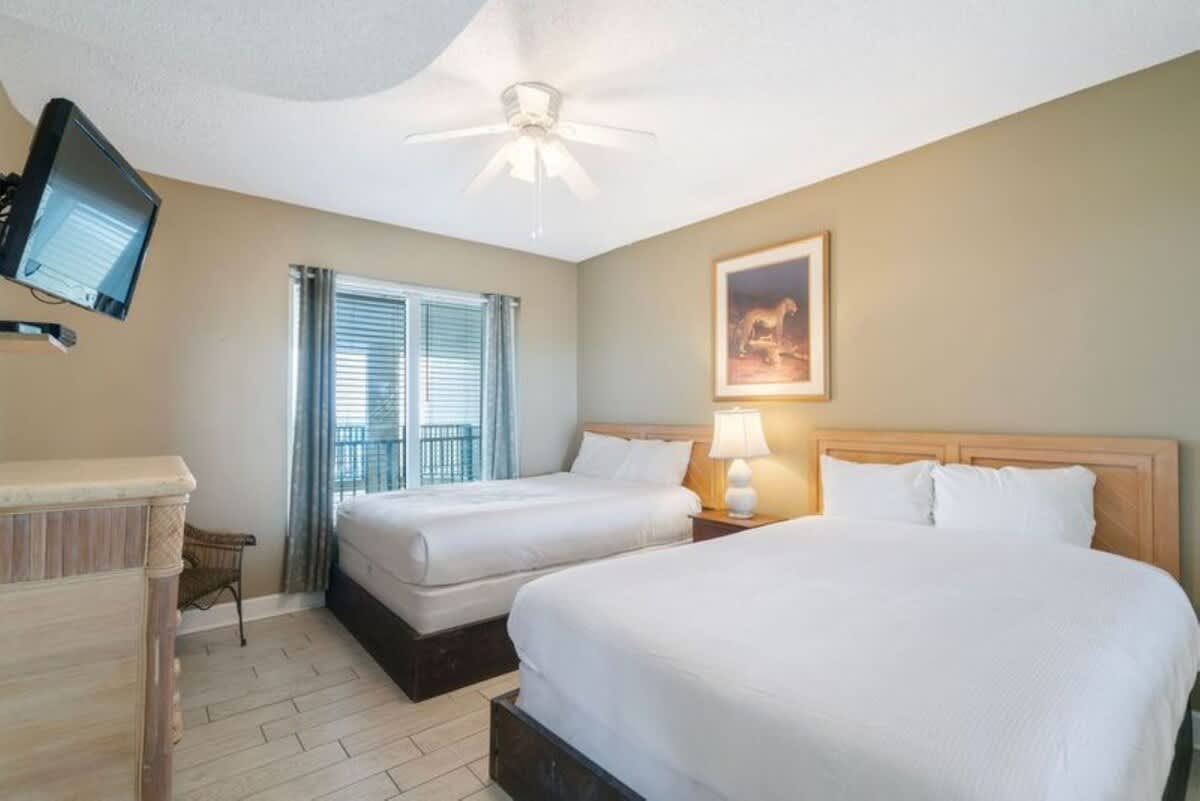 A bright bedroom with two queen beds, light wood headboards, and white bedding, featuring a ceiling fan and a wall-mounted TV. A sliding glass door leads to a balcony, offering natural light and a glimpse of the outdoors.