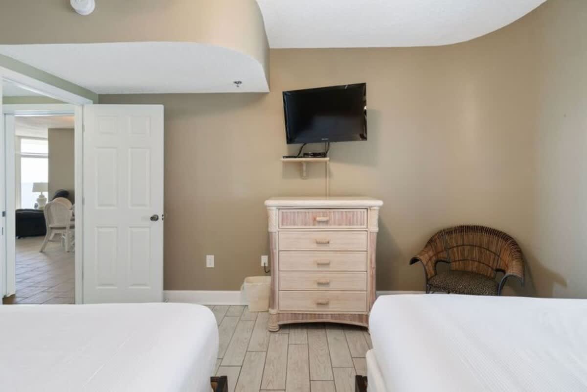 View of a bedroom with two queen beds and light wood-look tile flooring, featuring a light-colored dresser with a wall-mounted TV above it. A wicker chair with a patterned cushion adds a touch of texture, and an open doorway leads to another room.
