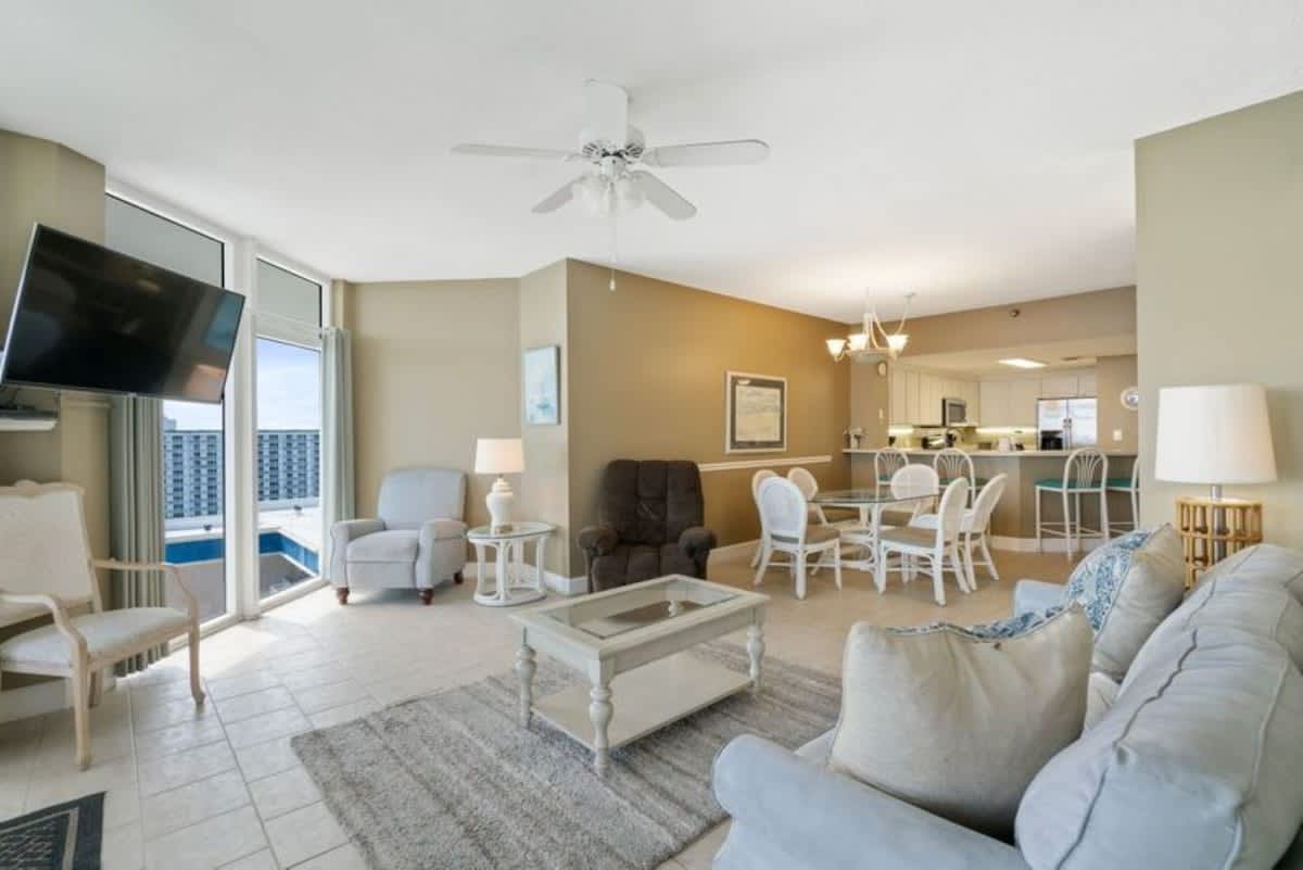 An expansive open-concept living area features a light sofa, a recliner, and a dining table with white chairs, all set on tiled floors, with large windows overlooking the ocean.