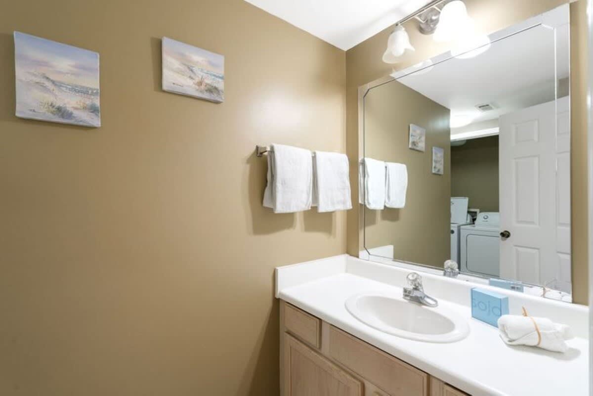 A modern bathroom vanity with a white countertop, single sink, and light wood cabinets. A large mirror reflects a laundry area with a washer and dryer, and the walls are painted a warm beige.