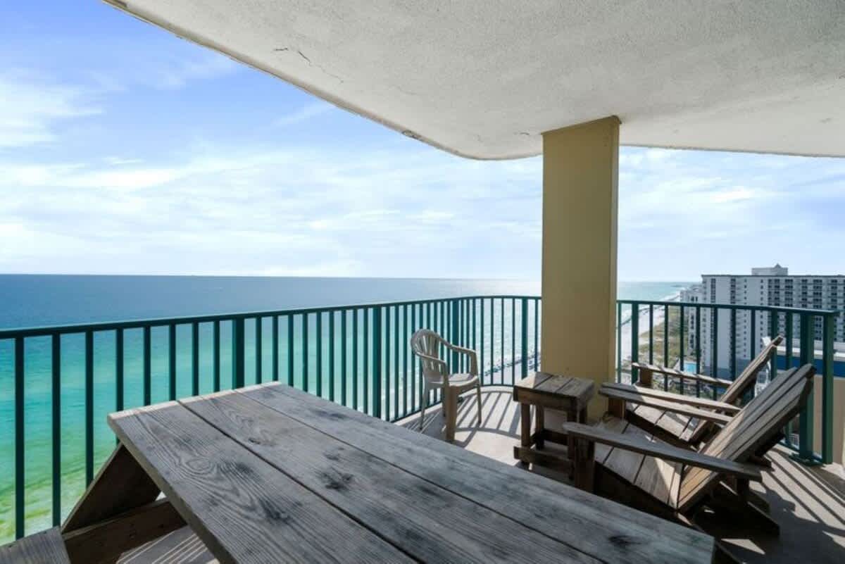 An expansive balcony offering panoramic ocean views, furnished with a large wooden picnic table and two wooden Adirondack chairs. The vibrant turquoise railing frames the stunning vista of the sea and distant coastline.