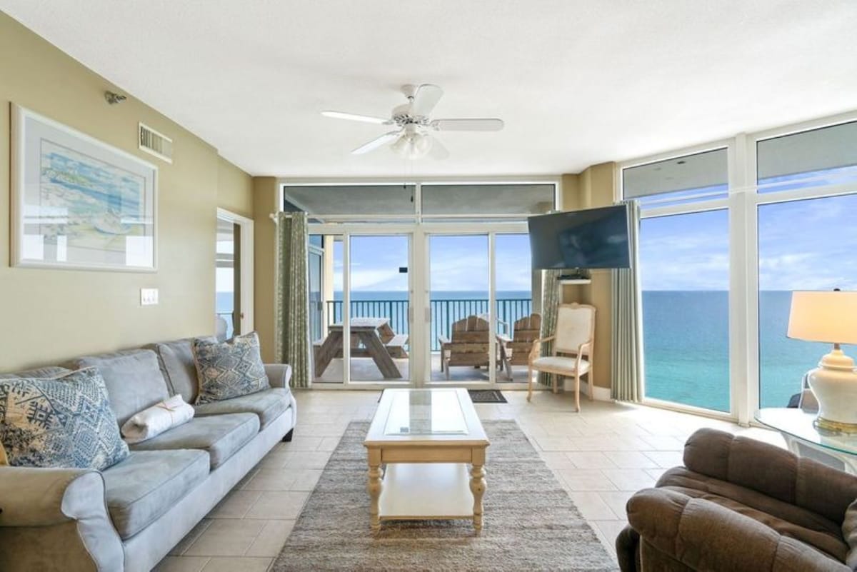 This bright living room offers stunning ocean views through expansive windows and sliding glass doors leading to a balcony, furnished with a light sofa, coffee table, and a wall-mounted TV.