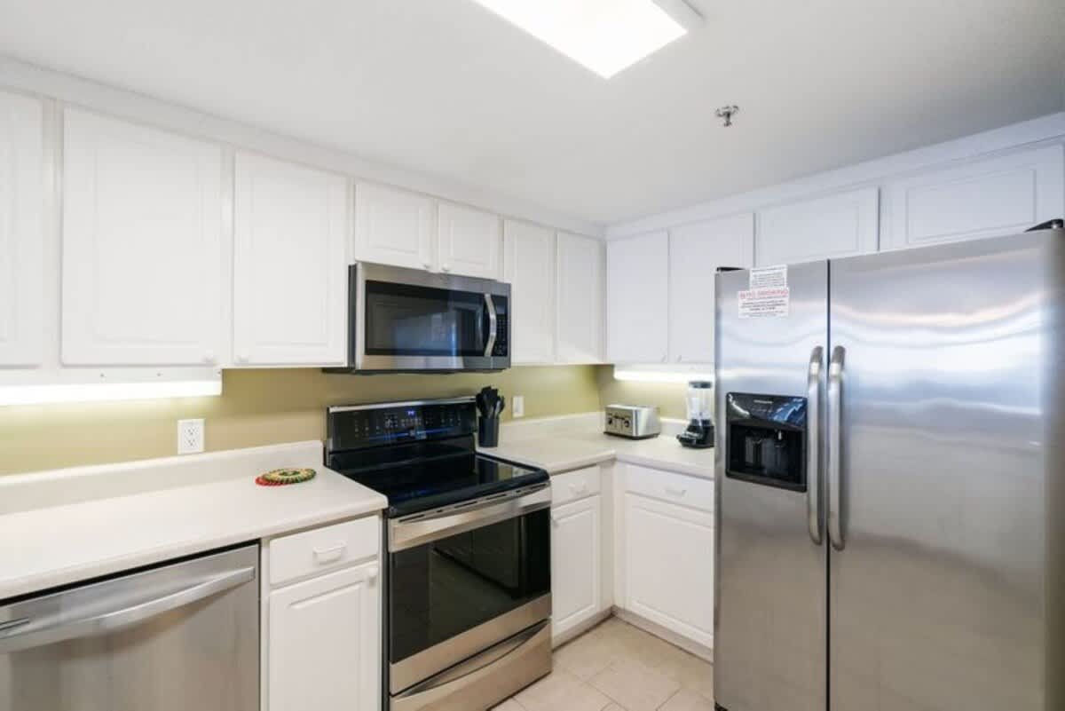 A modern kitchen features white upper and lower cabinets, light countertops, and stainless steel appliances including a refrigerator, microwave, dishwasher, and electric range. Under-cabinet lighting illuminates the backsplash.
