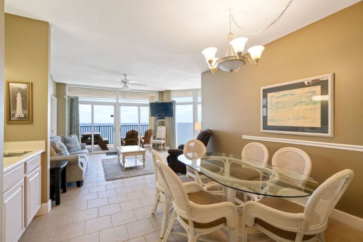 The dining area features a glass-top oval table with six white wicker chairs, illuminated by a chandelier, and is open to the living room with ocean views in the background.