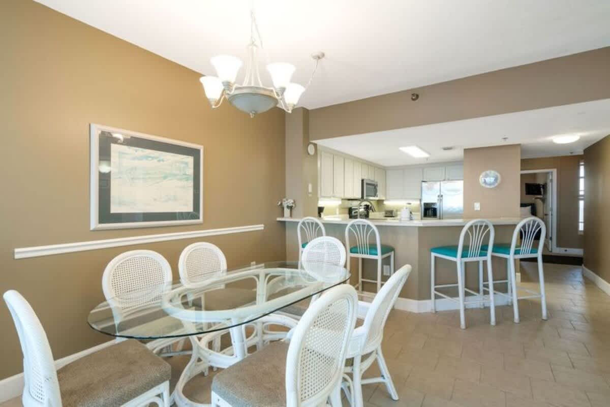 The dining room features an oval glass-top table with six white wicker chairs, a chandelier, and a framed artwork on the wall, adjacent to a breakfast bar with teal-seated stools.