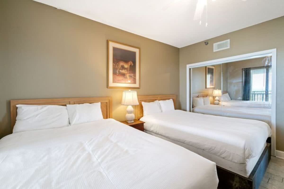 A cozy bedroom featuring two queen-sized beds with white linens and light wood headboards. A nightstand with a white ceramic lamp sits between the beds, and a large mirrored closet reflects the room.