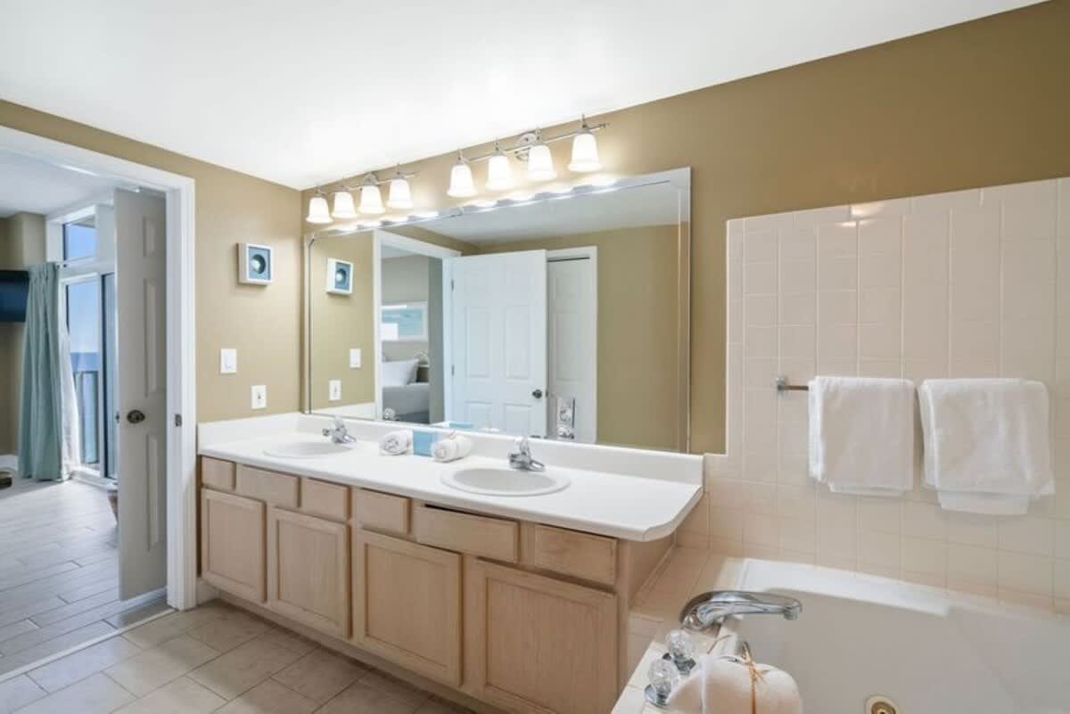 This master bathroom features a light wood double vanity with two sinks, a large mirror, and a jetted bathtub. The room has light tan walls and tile flooring, with an open doorway leading to the bedroom.