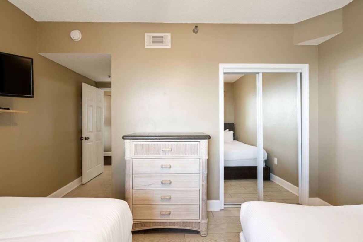A bedroom with two beds and light wood-look tile flooring, showing a light-colored dresser and a mirrored closet. An open doorway leads to a hallway, connecting to another room.