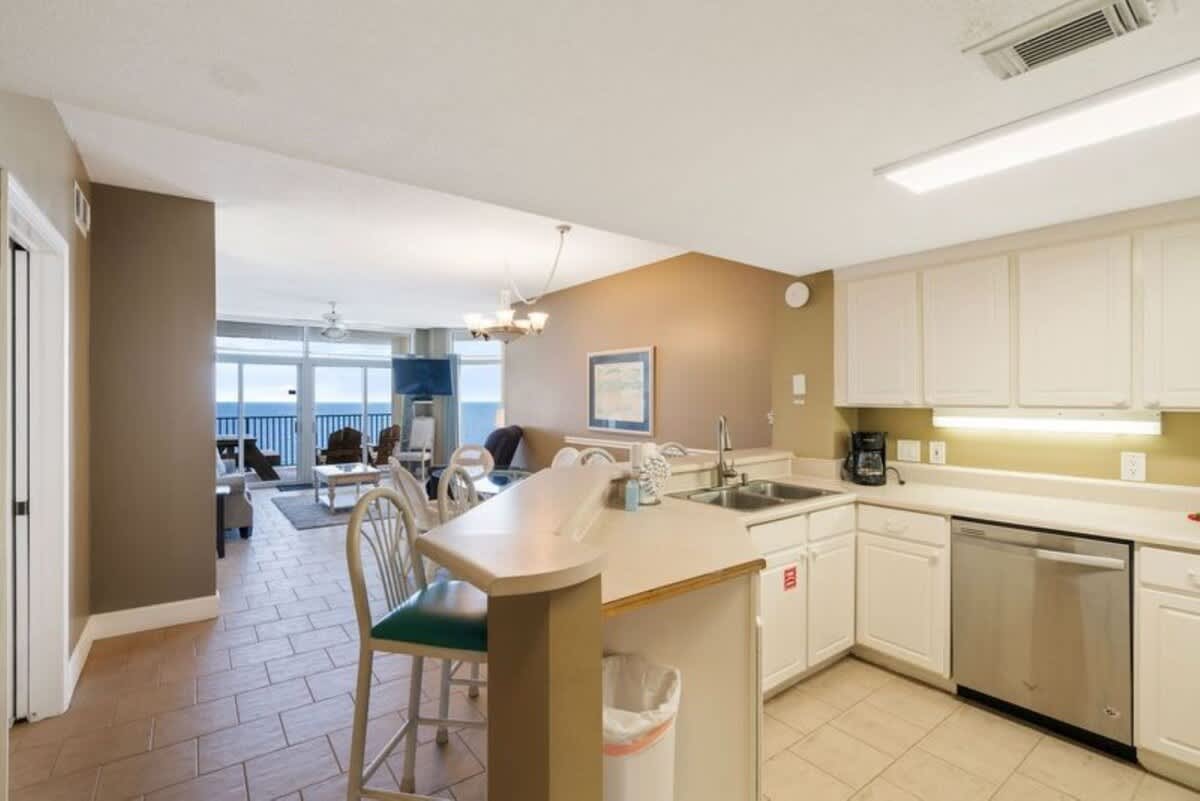 A spacious kitchen with white cabinets and light countertops includes a double sink and stainless steel dishwasher, featuring a breakfast bar that opens to the living and dining areas.