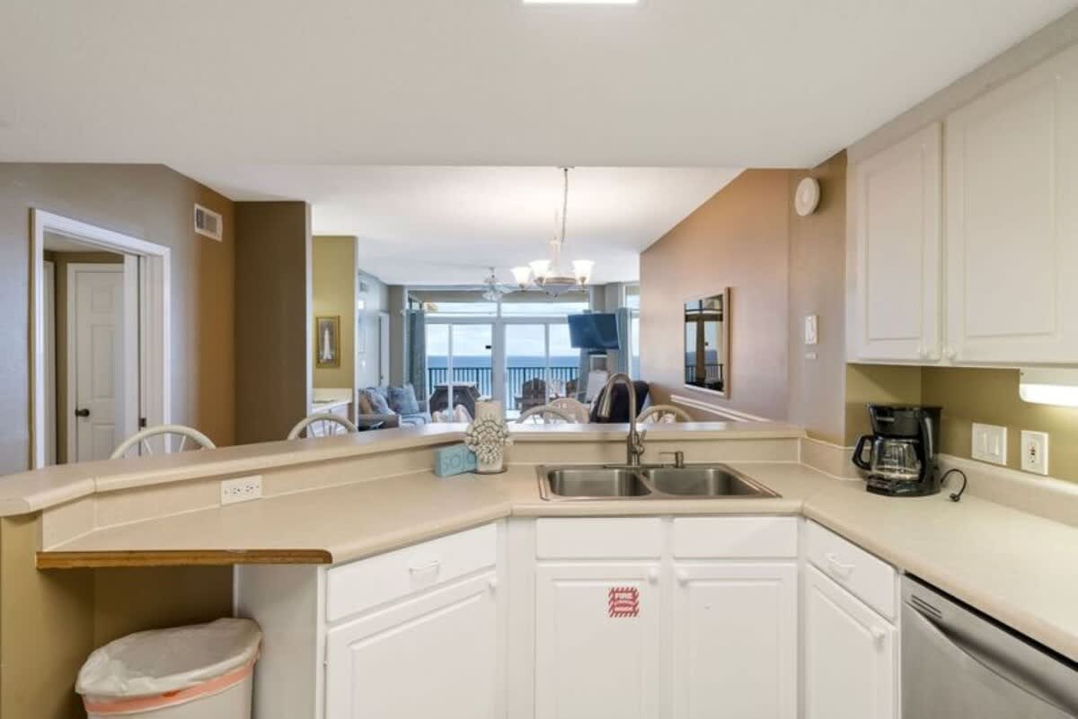 The kitchen showcases white cabinetry, light countertops, and a double sink, with a breakfast bar providing a pass-through view to the living room and ocean beyond.