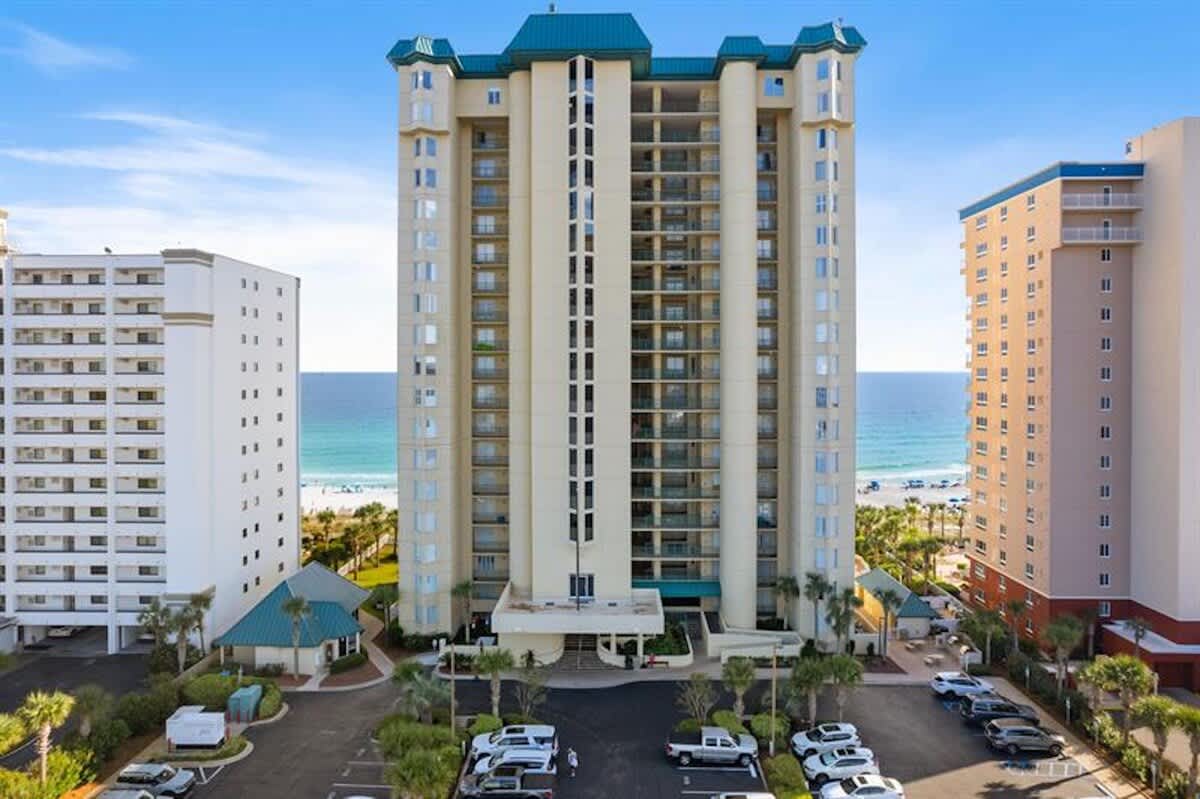 A tall, cream-colored condominium building with a distinctive blue roof stands prominently, featuring multiple balconies and a covered entrance. A large parking lot is in the foreground, with the ocean visible in the background.