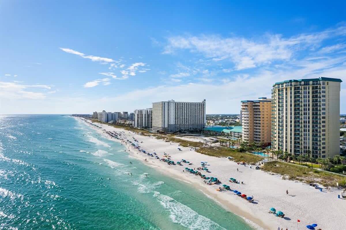 An expansive aerial view captures a long stretch of white sand beach lined with numerous beach umbrellas and people, with the clear turquoise ocean water gently lapping the shore. Several large condominium buildings are visible along the coastline under a bright blue sky.