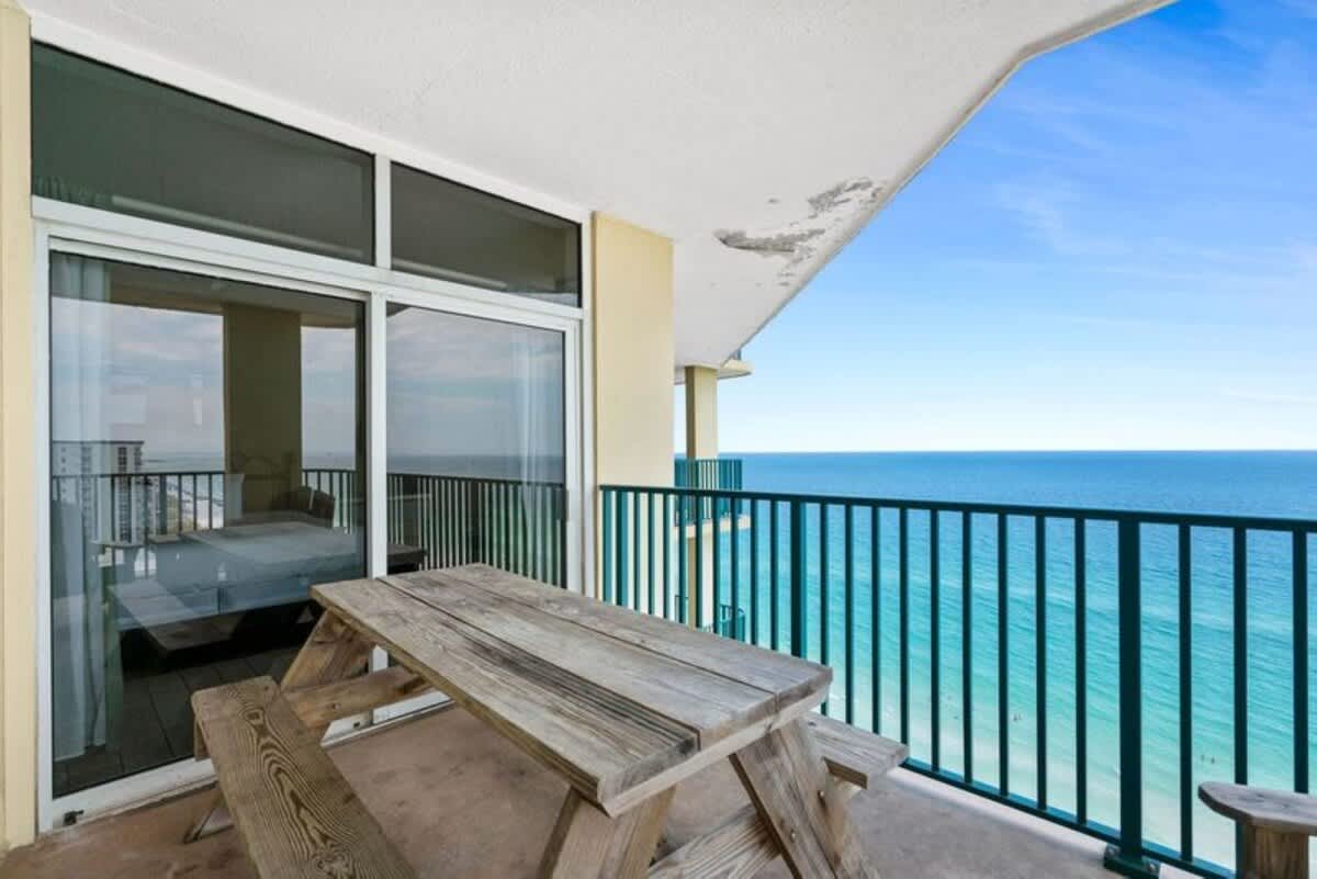 Wide-angle view of a concrete balcony with a large wooden picnic table, featuring sliding glass doors leading indoors and an unobstructed vista of the vibrant blue ocean and coastline.