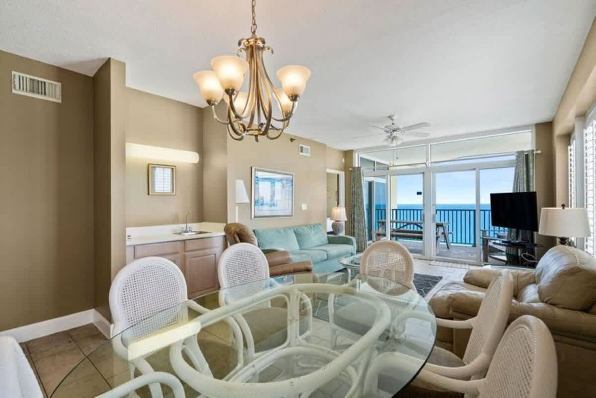 A view of the open-concept dining and living area, showcasing a glass dining table with white wicker chairs, a wet bar area, and the living room with ocean views in the background.