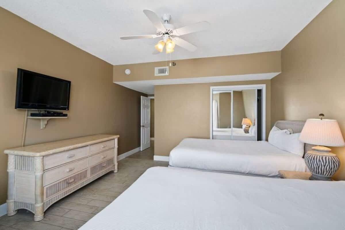 View of the second bedroom, showing two queen-size beds, a dresser with a wall-mounted TV, and a closet with mirrored sliding doors. The room has light-colored walls and a ceiling fan.