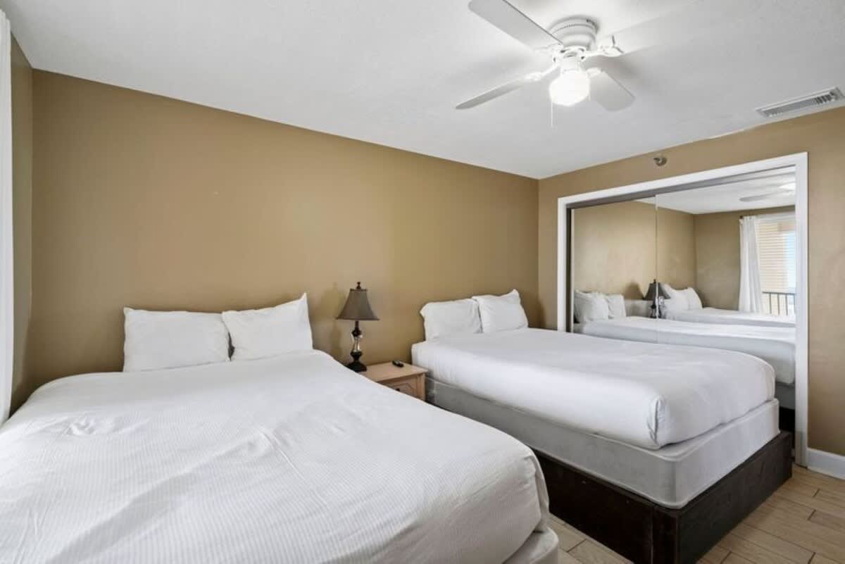 A bedroom featuring two queen-sized beds with crisp white bedding, a ceiling fan, and a large mirrored closet door reflecting the room. The walls are painted a warm tan.