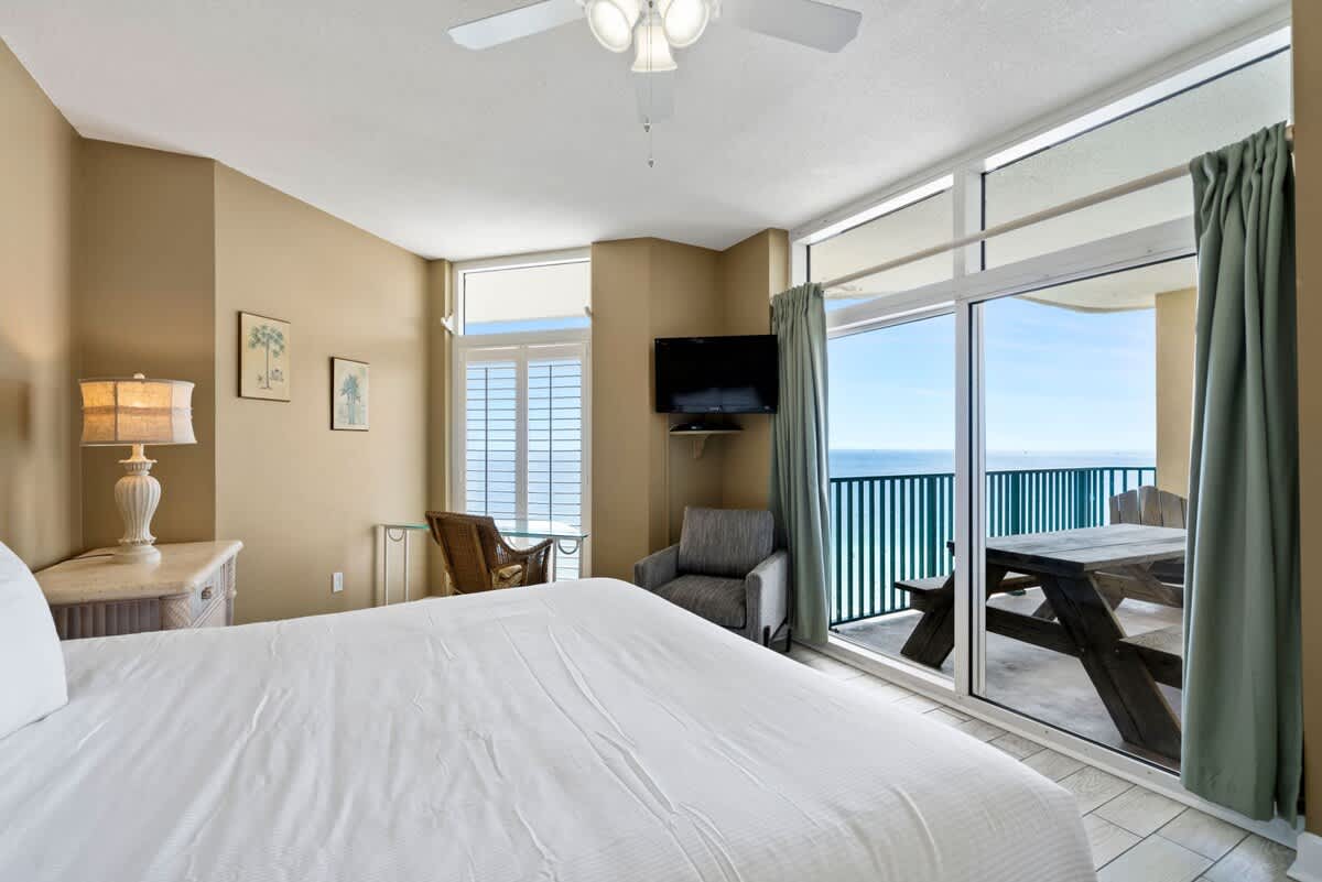 View of the bedroom from the bed, showcasing a large sliding glass door leading to a balcony with an ocean view. A wall-mounted TV, a gray armchair, and a small dining table are visible near the window.