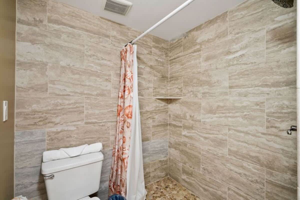 A bathroom with a white shower/tub combination, a toilet, and a vanity with a white sink and large mirror. White tiled walls contrast with tan painted walls.