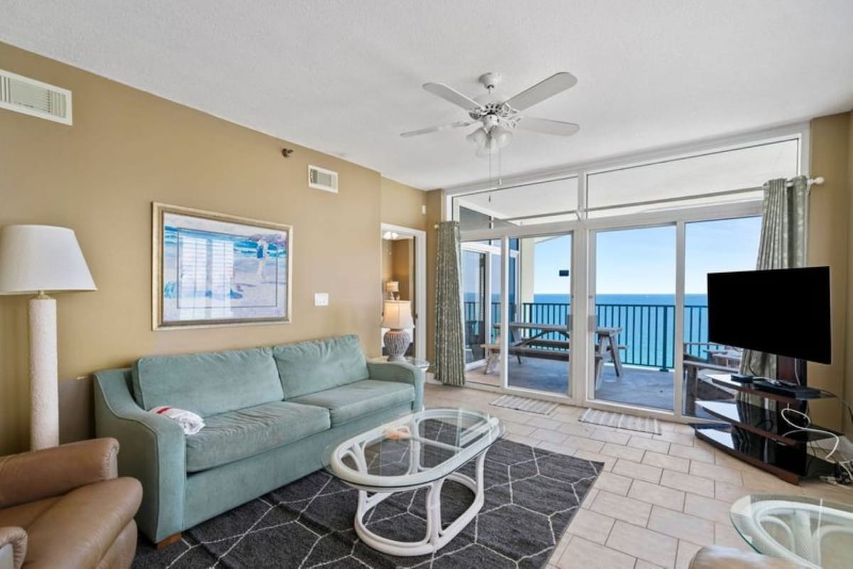 A slightly wider view of the living room, showing a light blue sofa, glass coffee table, and a TV stand, with large sliding glass doors providing expansive ocean views from the balcony.