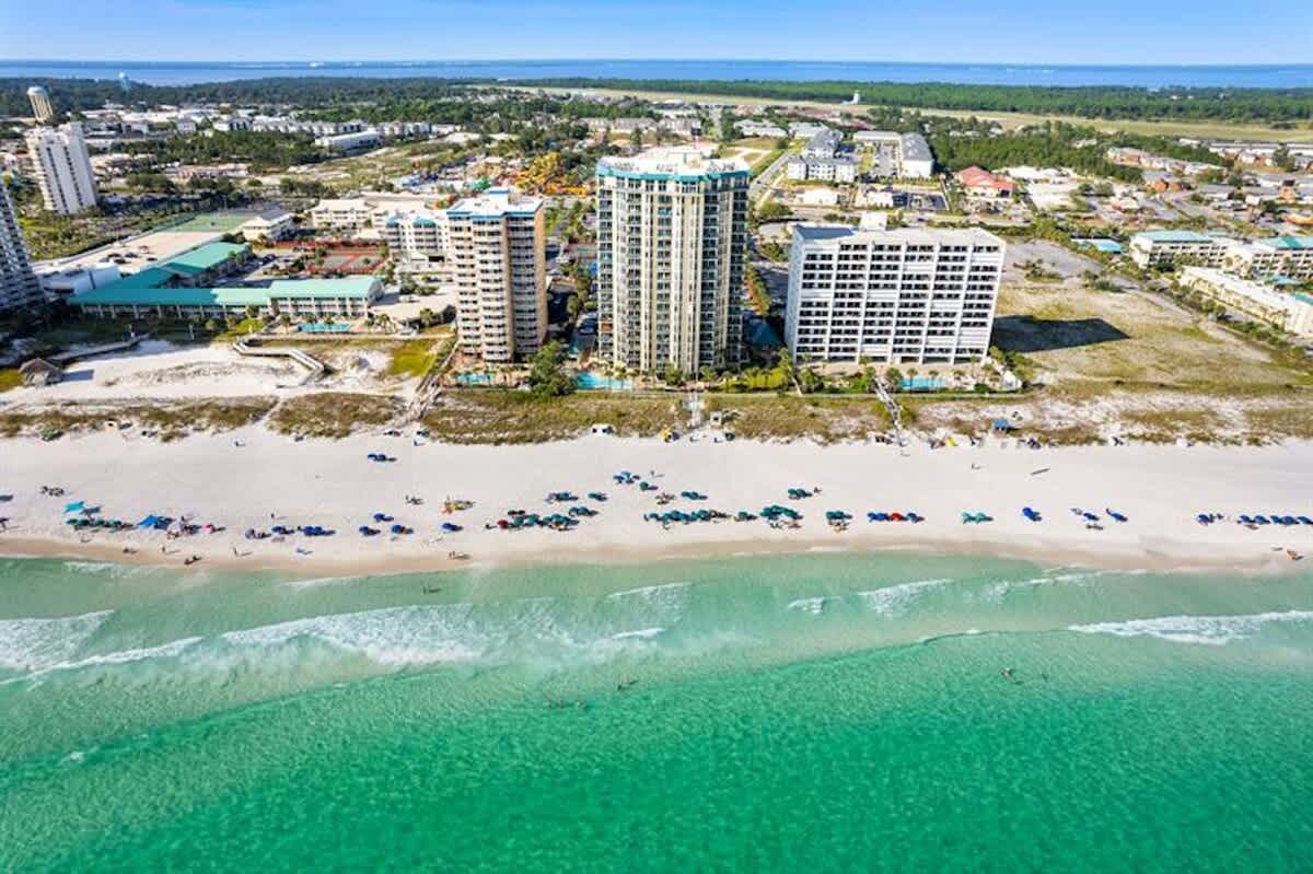 A high aerial perspective reveals a long, pristine white sand beach with turquoise ocean waters, dotted with beachgoers and umbrellas. Multiple large condominium complexes are visible along the coastline, with a vast expanse of land and water in the distance.