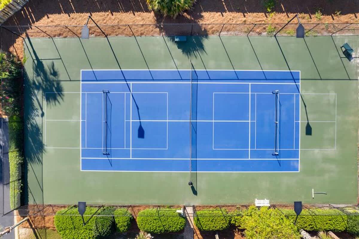 An aerial view showcases two adjacent blue and green tennis courts with white lines and nets, enclosed by a black fence and surrounded by green foliage and palm trees.