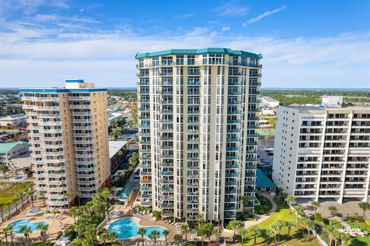 Aerial view of a tall, modern condominium building with a distinctive teal roof, surrounded by other high-rises, multiple swimming pools, and lush green landscaping, all set against a clear blue sky and distant ocean.