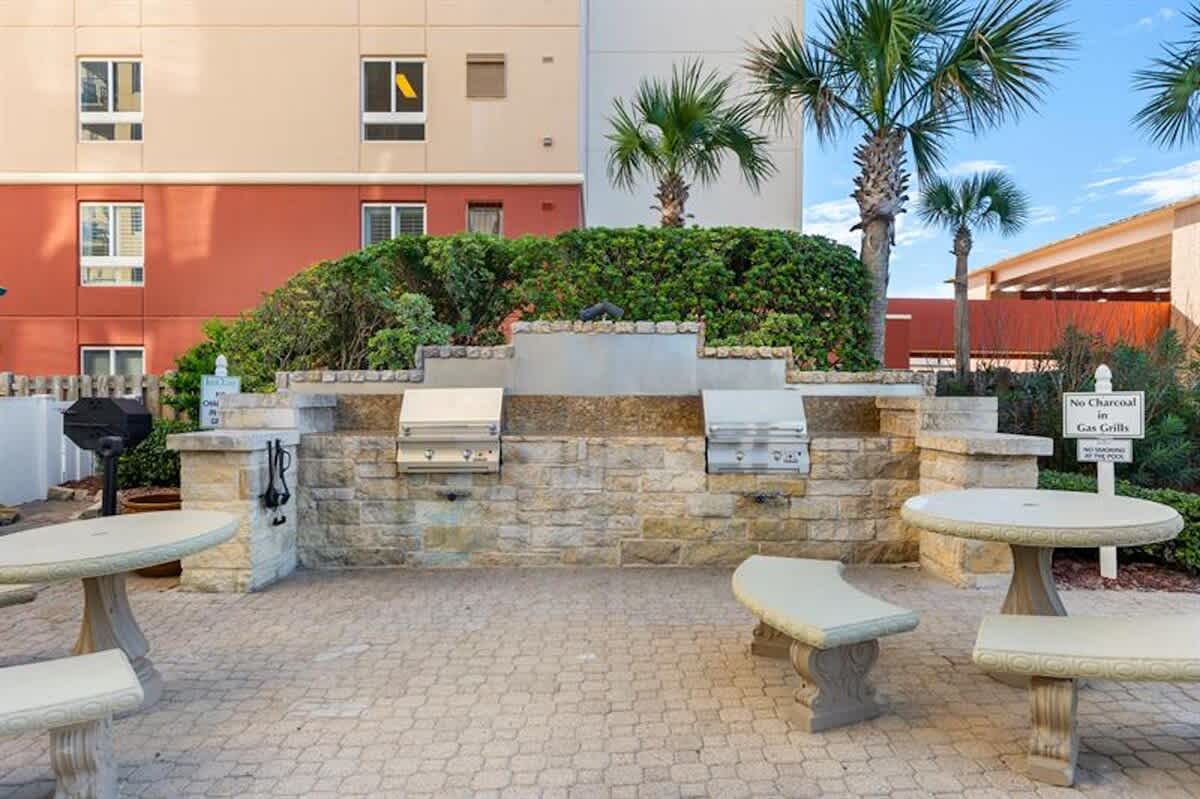 An outdoor grilling area features multiple built-in gas grills set into a stone and concrete counter, surrounded by paved patio flooring and several round tables with benches. Palm trees and lush green bushes are visible in the background against a building.