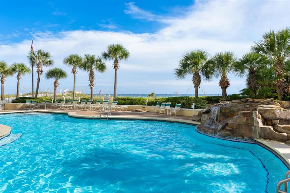 A large, irregularly shaped outdoor swimming pool with clear blue water, surrounded by lounge chairs and lush palm trees, featuring a rock waterfall.
