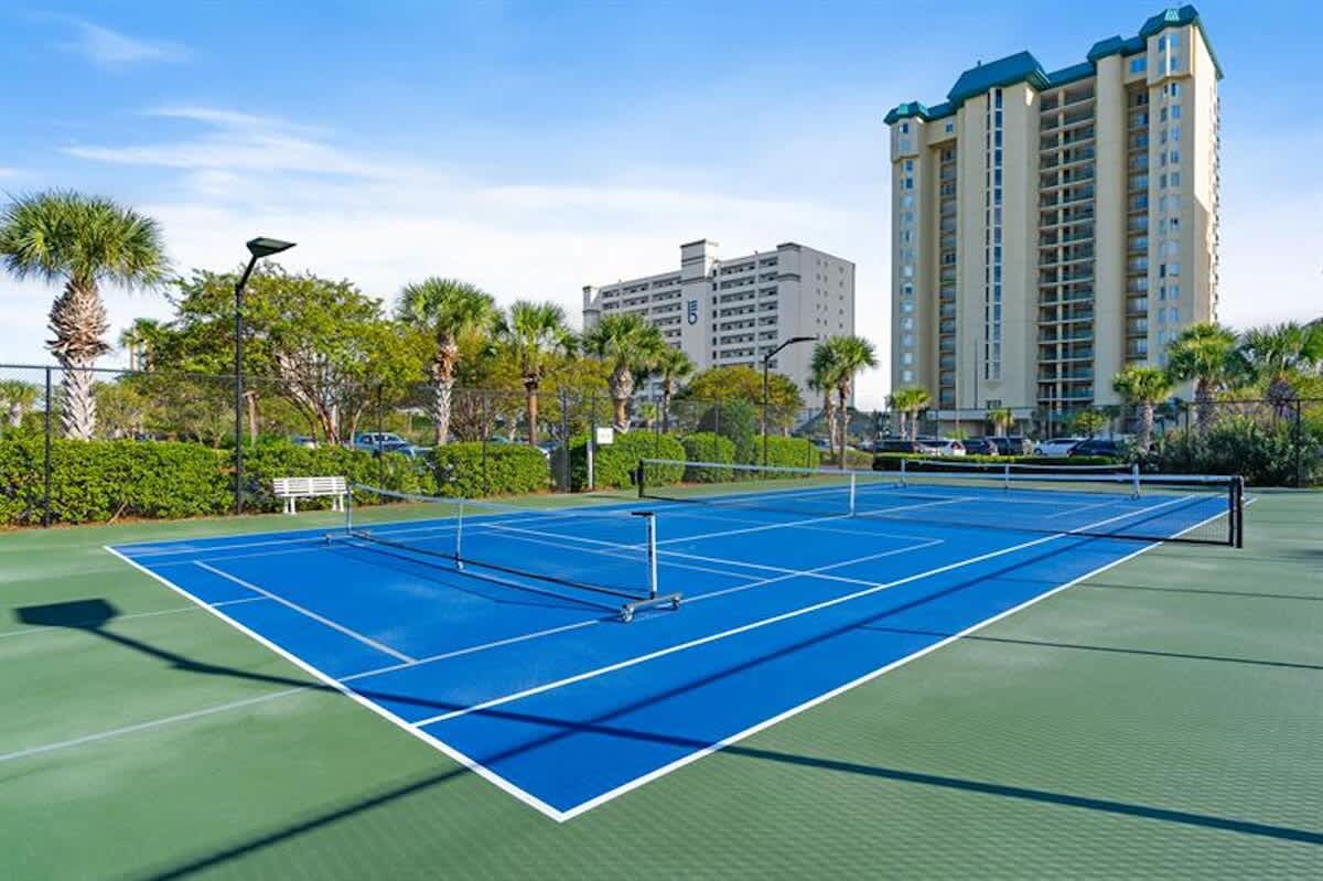 A wide shot of a bright blue and green tennis court, featuring a net and white boundary lines, surrounded by lush green bushes and palm trees, with tall resort buildings in the distance.