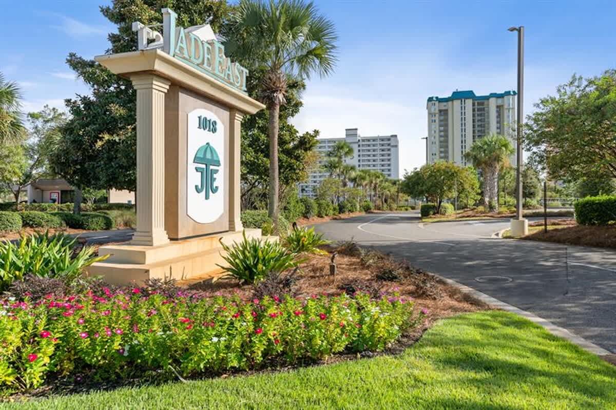 The entrance to the property is marked by a large sign displaying "JADE EAST" and "1018", surrounded by colorful flowers and lush landscaping. A paved road leads towards the condominium buildings in the distance under a clear sky.