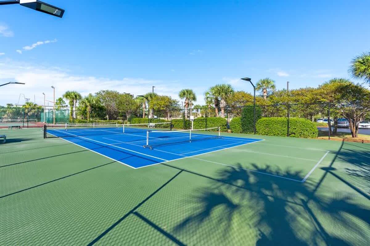 Two side-by-side blue and green tennis courts are visible, separated by nets, surrounded by green landscaping and tall light poles, with a clear blue sky overhead.