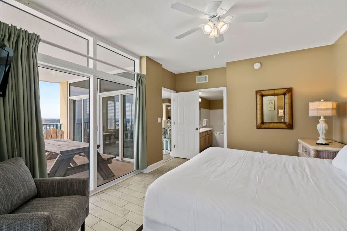 Bedroom view towards the entrance, revealing a doorway to an en-suite bathroom. The room features a king-size bed, a gray armchair, and a large sliding glass door opening to a balcony with an ocean view.