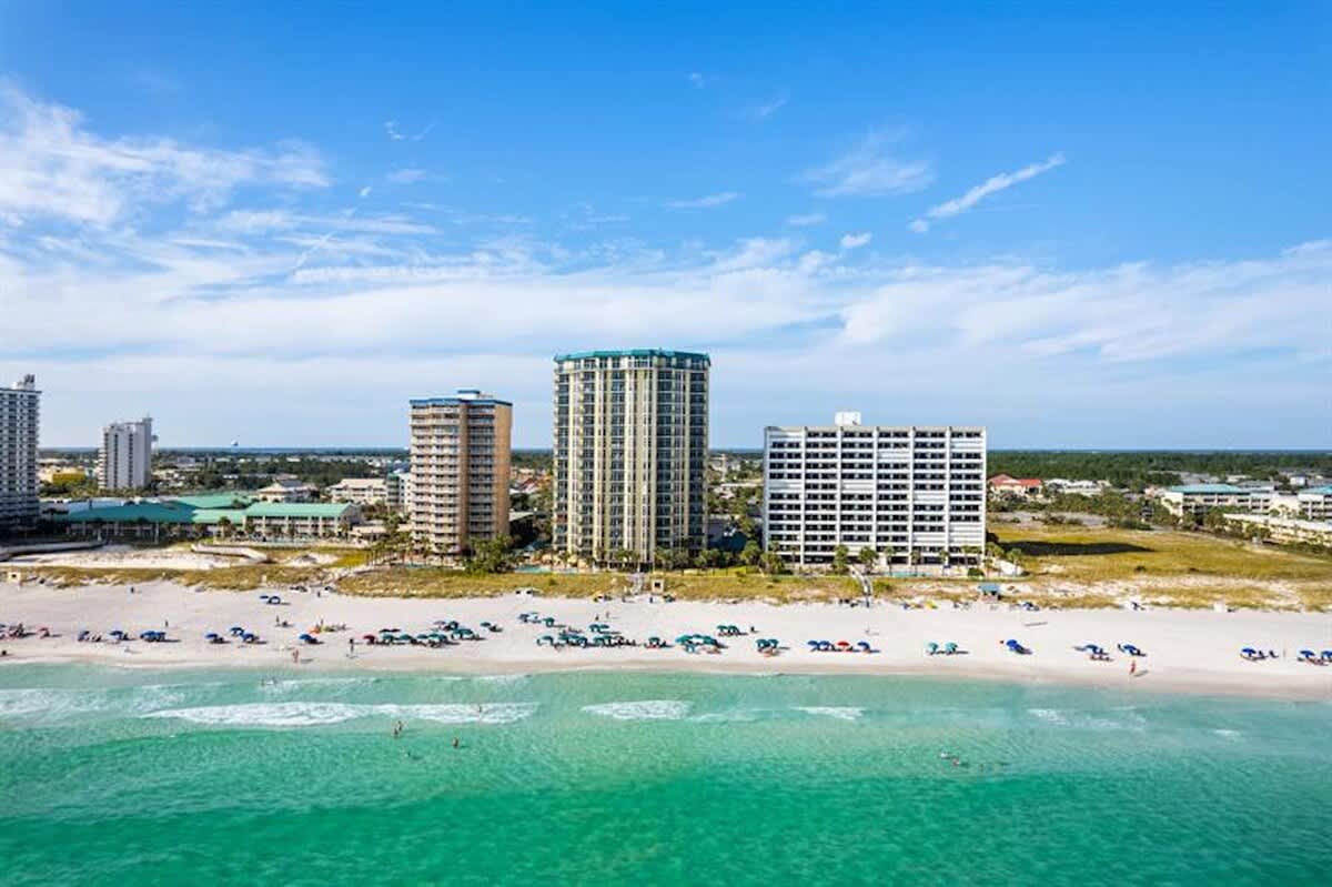 A wide aerial shot shows a vibrant white sand beach with many colorful umbrellas and people enjoying the clear turquoise ocean. In the background, several tall condominium buildings stand along the coast, with a clear blue sky above.