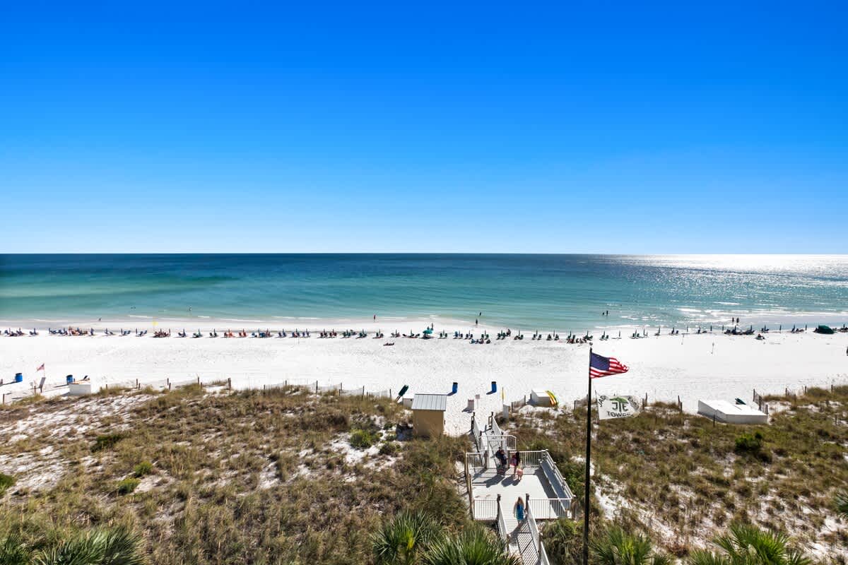 A breathtaking panoramic view of a wide, white sand beach lined with umbrellas and the vast, sparkling turquoise ocean under a clear blue sky, seen from the property's balcony.