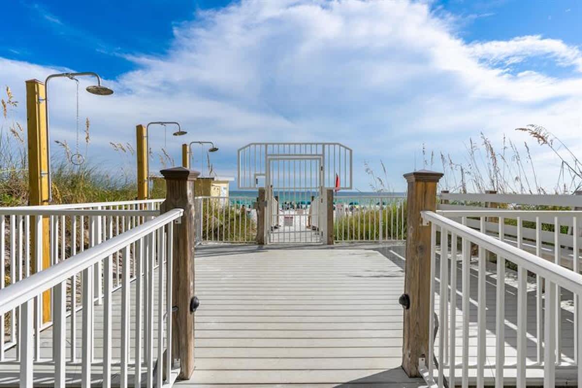 A wooden boardwalk leads towards the beach, featuring white railings and outdoor showers on either side, with dune grasses and the ocean visible through a gate in the distance.