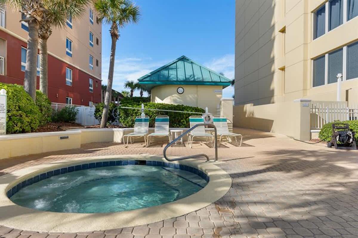 A bubbling circular hot tub is set into a brick paver patio, surrounded by lounge chairs and lush green landscaping, with tall buildings and palm trees under a clear blue sky.