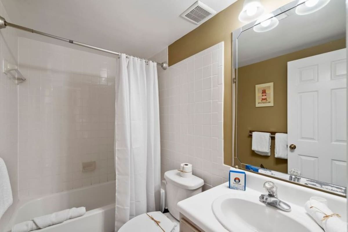 A bathroom with a white shower/tub combination, a toilet, and a vanity with a white sink and large mirror. White tiled walls contrast with tan painted walls.