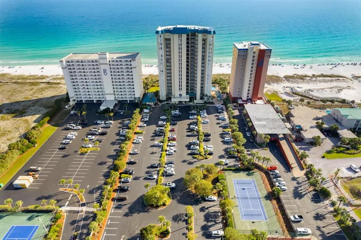An aerial view of the condominium complex, showcasing multiple high-rise buildings, a large parking lot, tennis courts, and its prime beachfront location.